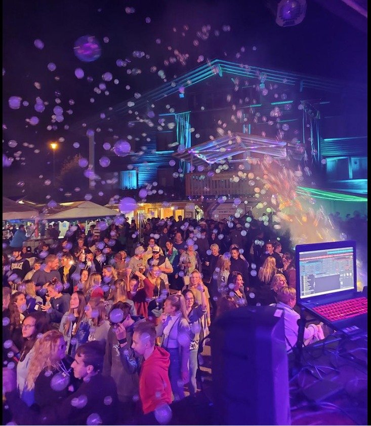 Menschen tanzen und feiern auf einer Party im Freien bei Nacht, mit bunten Lichter und einer Diskokugel, im Vordergrund ein DJ-Pult mit Laptop.
