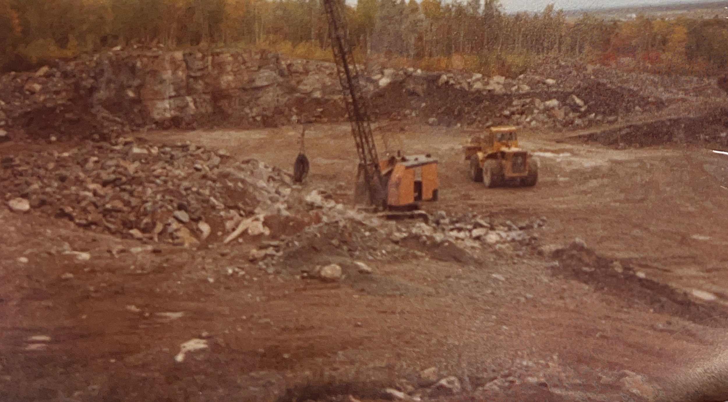 Vue d'une carrière avec des machines de construction, notamment une pelleteuse et un camion chargeur, entourées de piles de pierres et de terre, avec une forêt en arrière-plan.