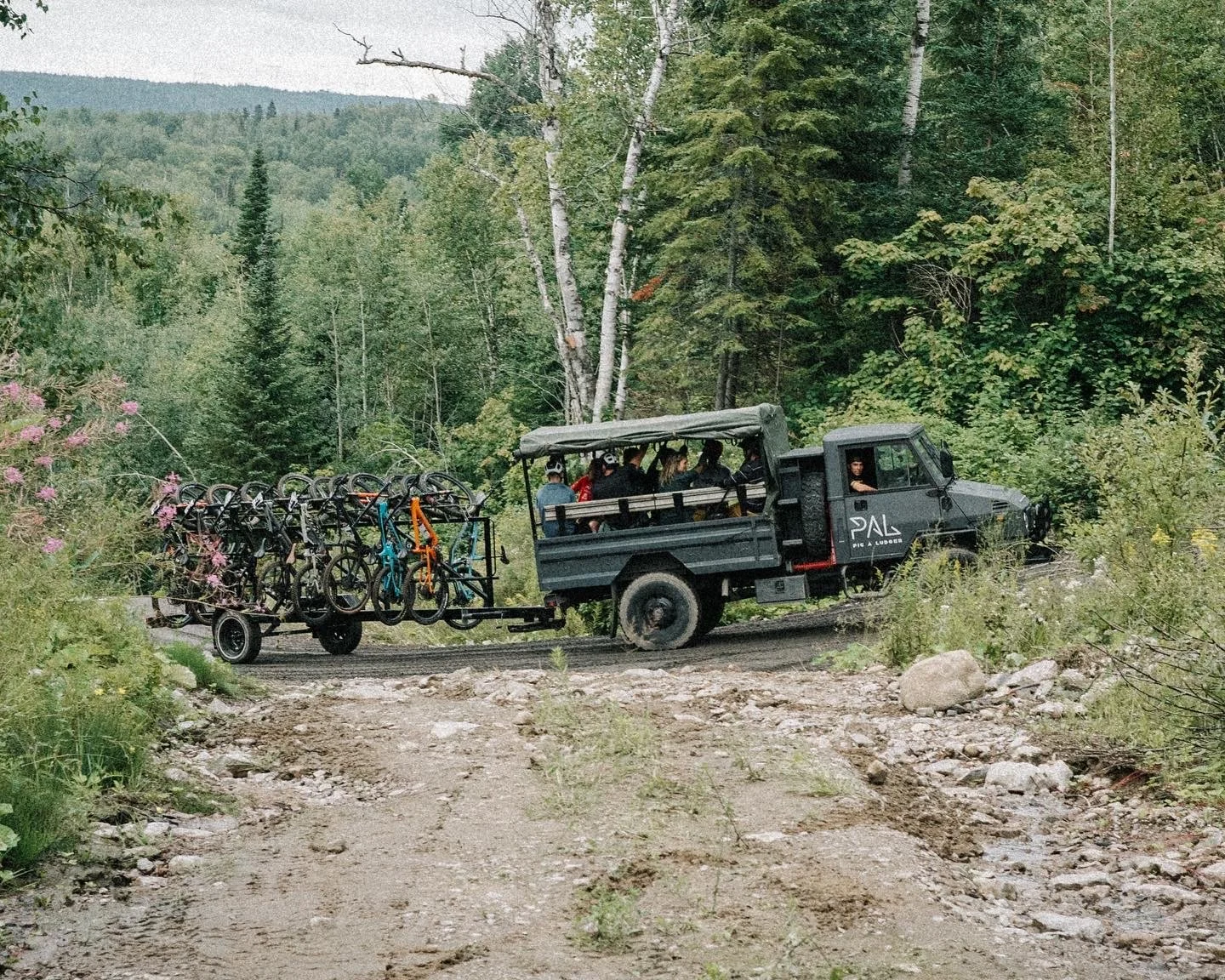 Un véhicule tout-terrain transporte des vélos sur une remorque dans une forêt pittoresque, sous un ciel gris. Plusieurs personnes sont assises à l'intérieur du véhicule qui porte l'inscription "PAL" sur le côté.