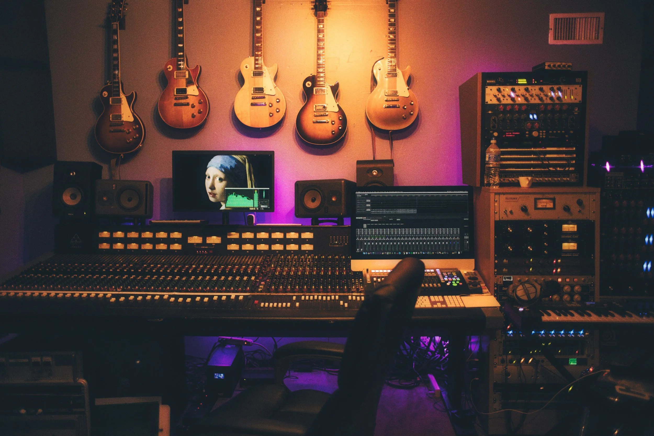 A music recording studio with wall-mounted electric guitars, a computer monitor showing a woman painted in art style, a mixing console, studio speakers, and various audio equipment illuminated with colorful lights.