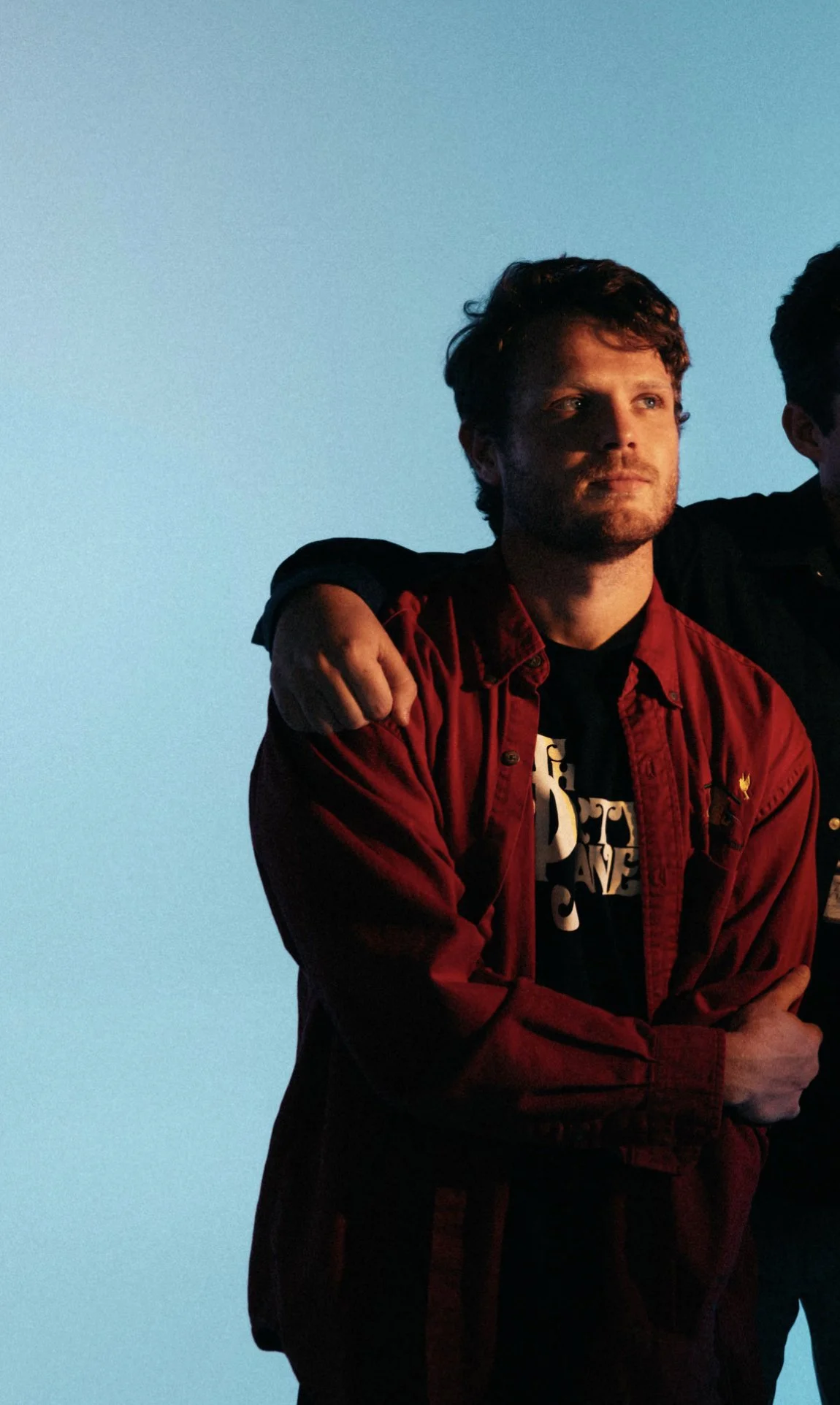 A man with curly brown hair and a beard, wearing a red jacket over a black graphic T-shirt, stands with his arms crossed against a blue sky background.