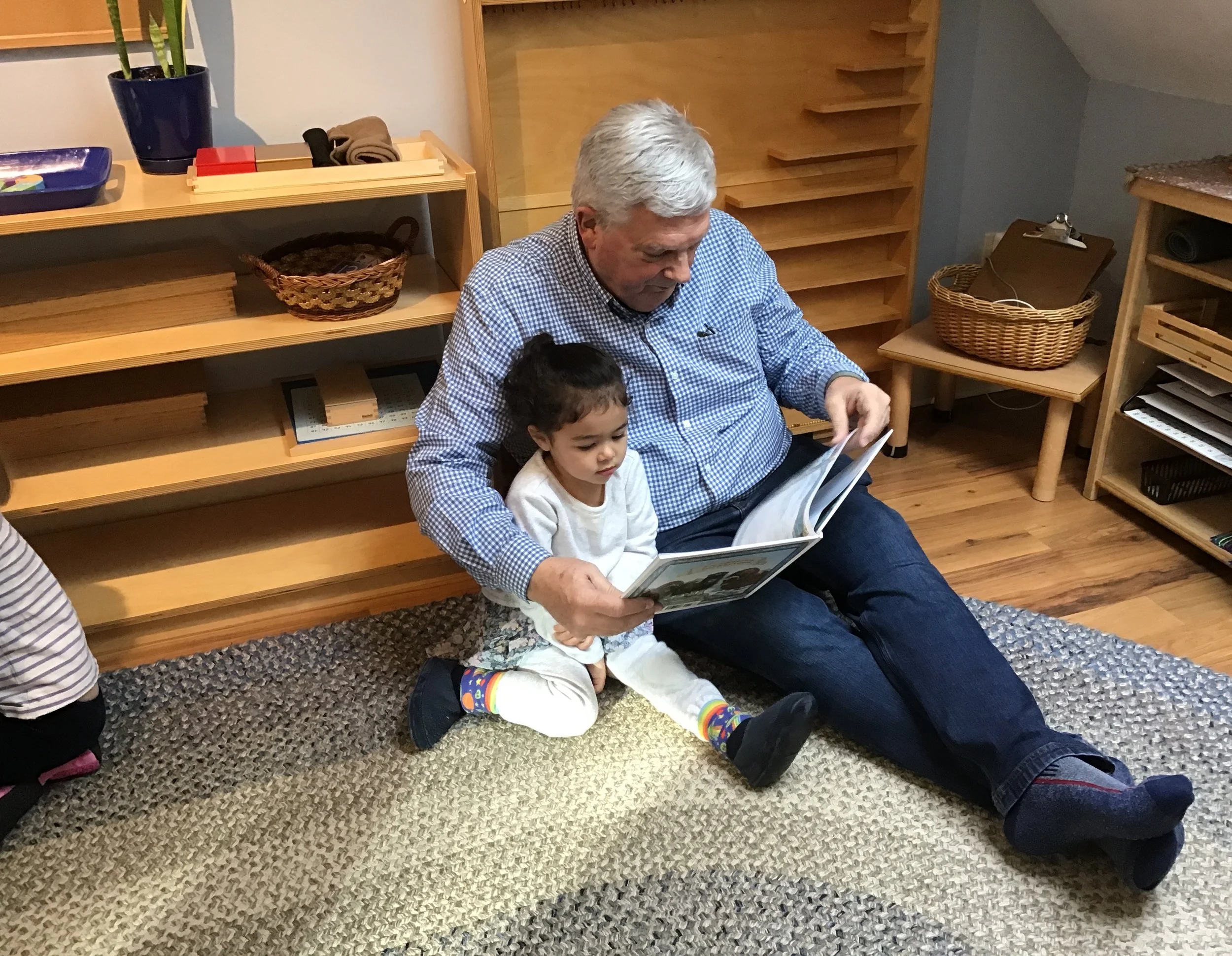A grandfather and his grandchild sitting on the floor, reading a book together in a cozy room with wooden shelves and a basket.