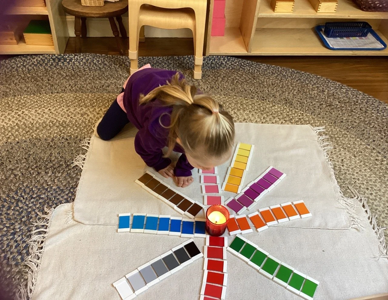 A young girl with blonde hair on a white cloth, arranging color swatches in a circular pattern around a lit red candle on a woven rug.