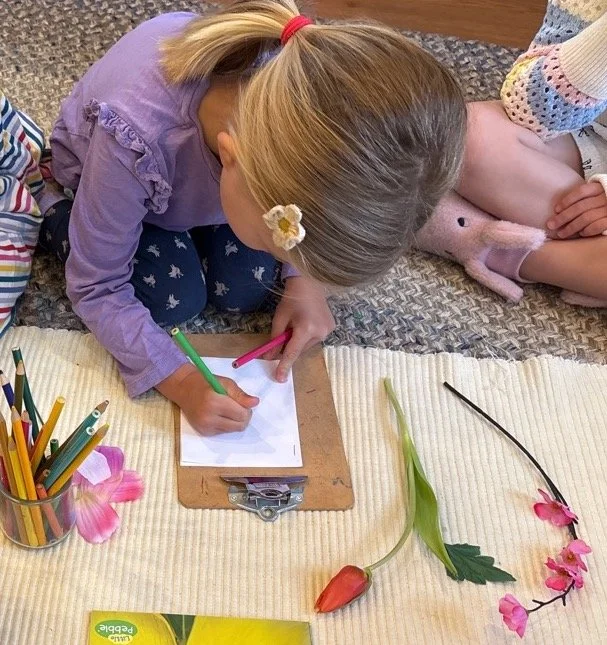 A young girl with a white flower hair clip is drawing with colored pencils on a clipboard resting on a table covered with a yellow cloth. There are flowers and a container of colored pencils on the table, with part of a person sitting next to her and a pink stuffed animal nearby.