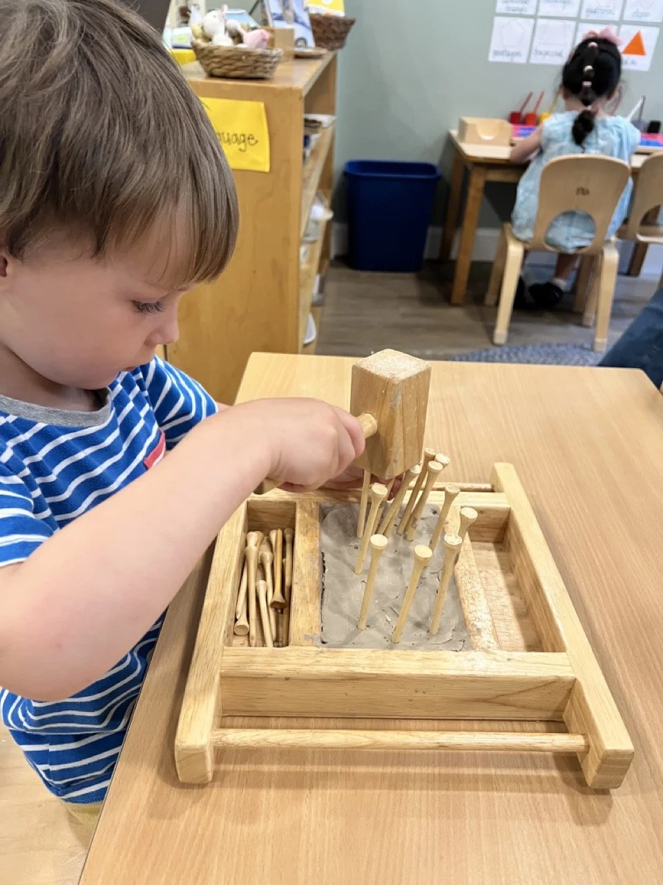 A young boy in a blue striped shirt playing with a wooden sandbox and small wooden pegs in a classroom.