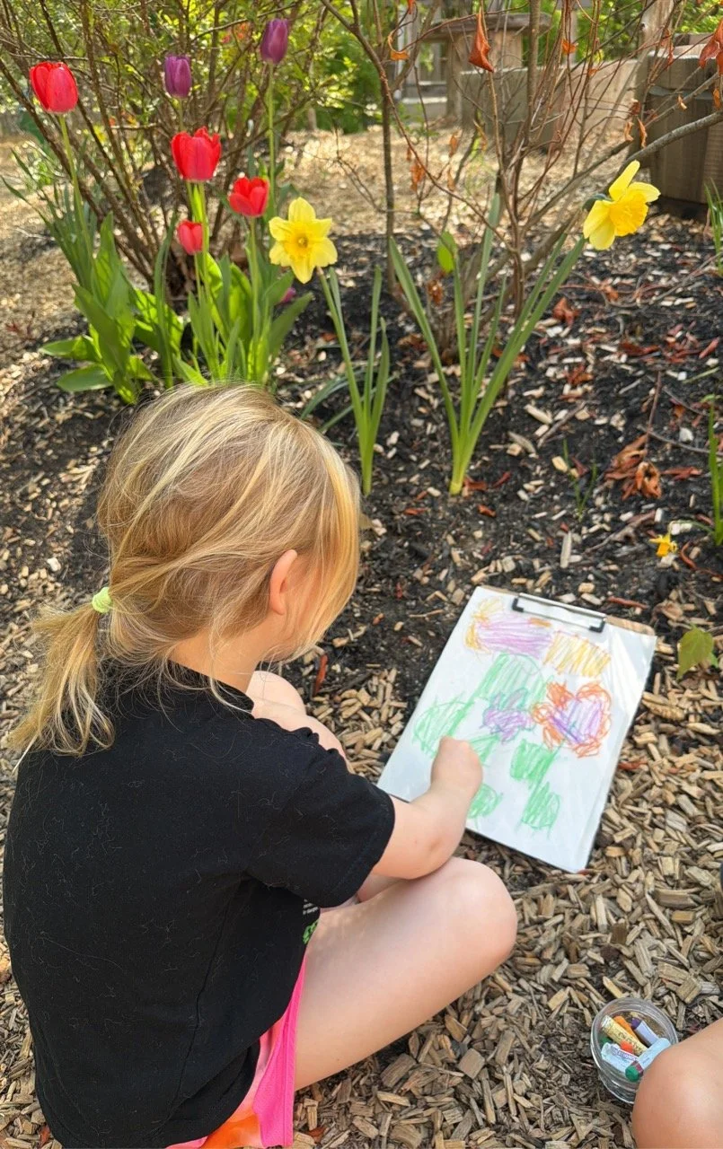 A young girl with blonde hair tied in a ponytail, sitting outdoors among tulips and daffodils, drawing on a clipboard.