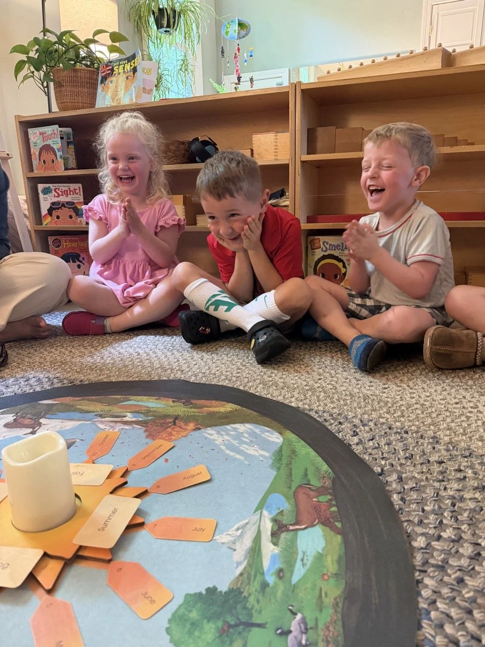 Three children sitting on a carpet and laughing, with a bookshelf behind them and a colorful educational game on the floor in front of them.