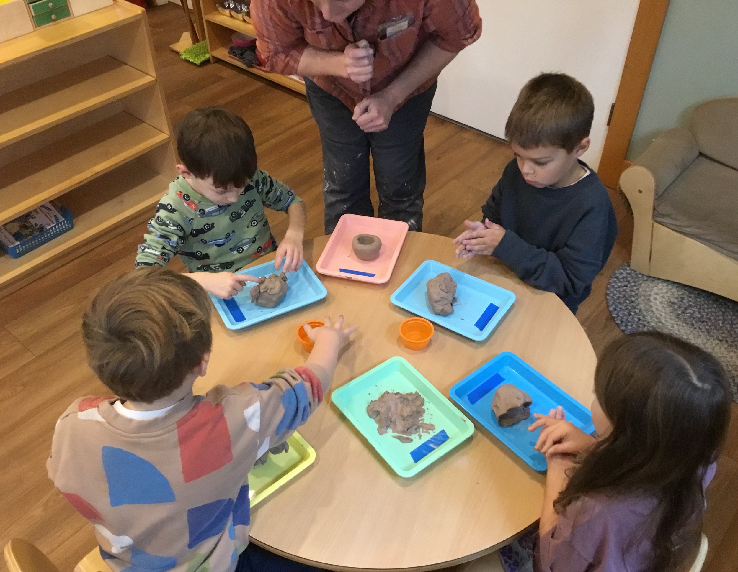 Five children sitting and standing around a wooden table with trays of clay, working on clay projects, under the supervision of an adult.
