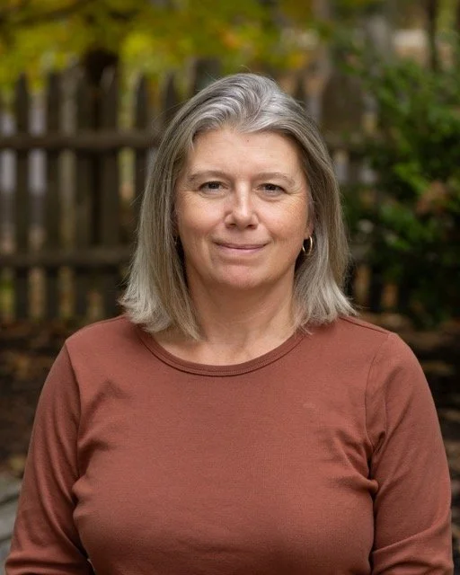 A woman with shoulder-length gray hair and earrings, wearing a brown shirt, standing outdoors in front of a wooden fence and trees.