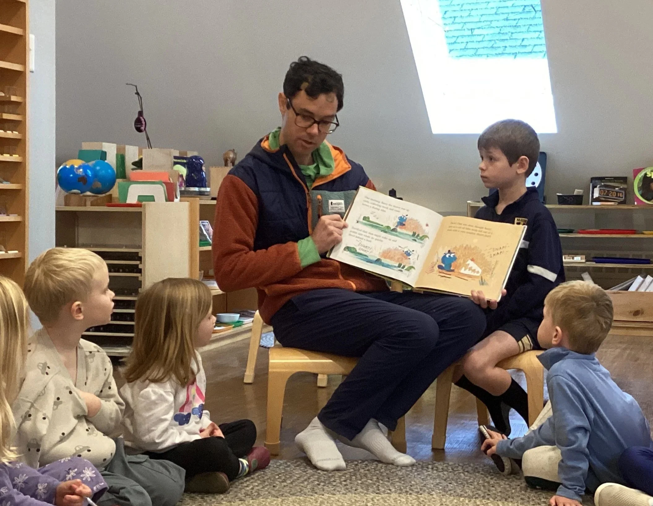 A man reading a children's book to a group of kids seated on the floor in a classroom or library.