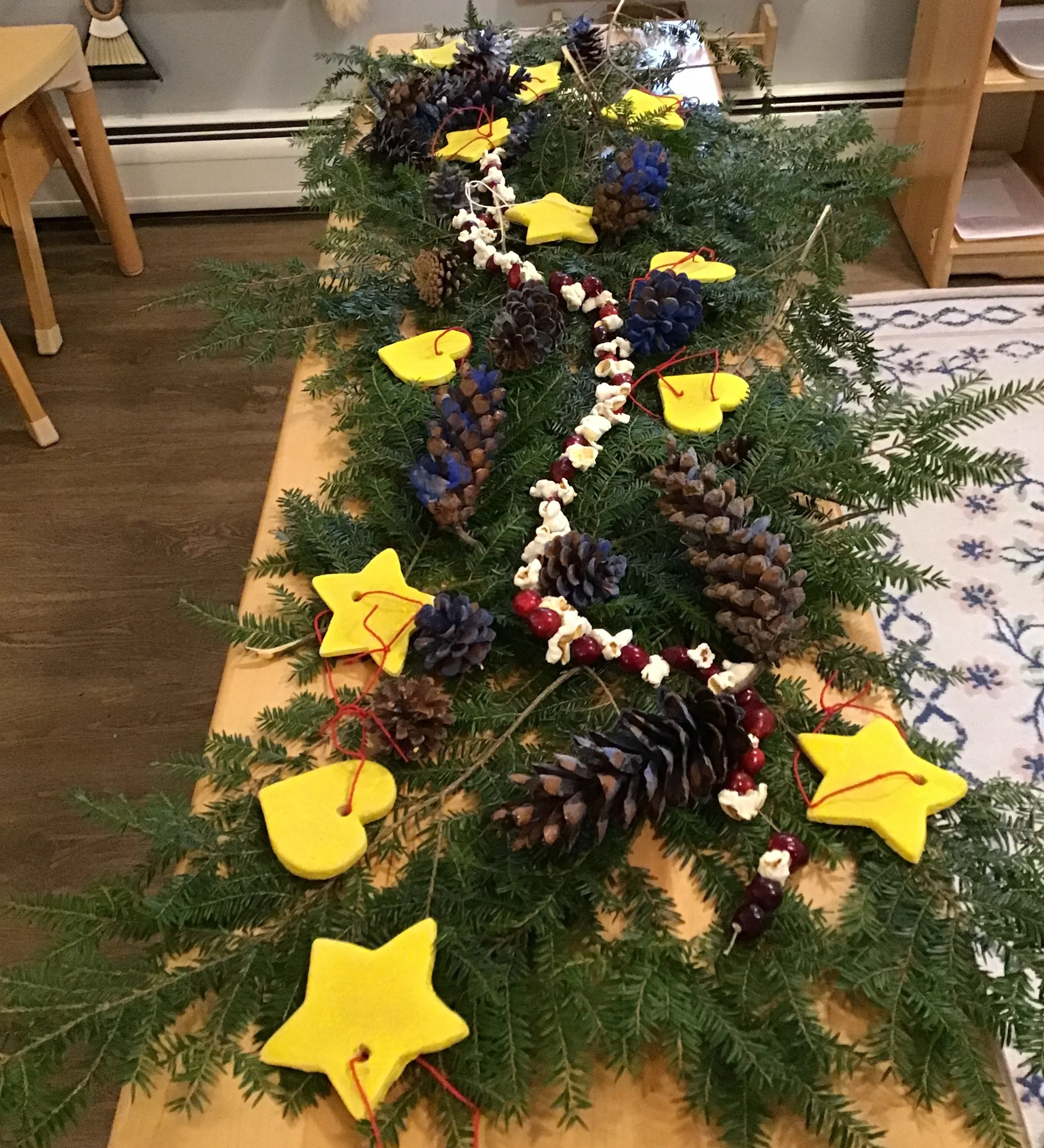 A collection of pine boughs, decorated for the Solstice, including yellow star-shaped ornaments, pine cones, blue berries, and white popcorn garland, all on a wooden table in a cozy indoor setting.