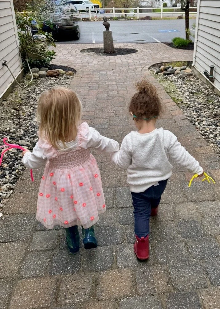 Two young girls walking hand in hand on a brick pathway, wearing rain boots, with a small sculpture on a post in the background.