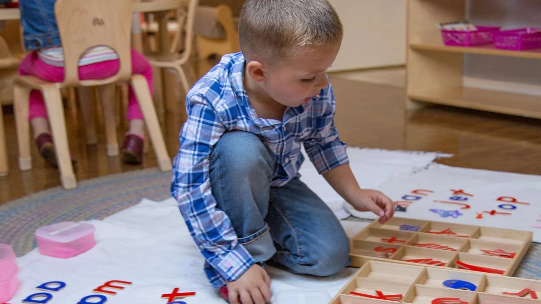 A young boy kneels on a blanket, playing with wooden letter tiles and letter boxes, in a classroom or playroom setting.