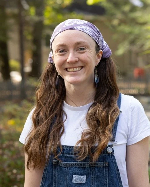 A woman with long brown hair, wearing a purple bandana, white t-shirt, and denim overalls, smiling outdoors in a park-like setting.