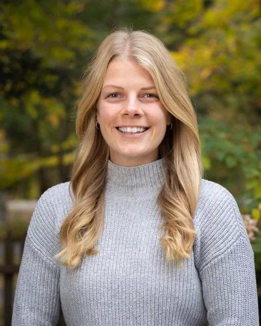 A young woman with blonde wavy hair, smiling at the camera, wearing a gray turtleneck sweater, with a background of autumn foliage.
