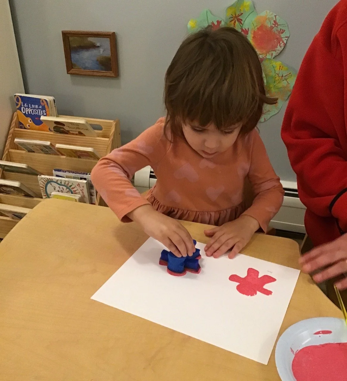 A young girl with brown hair wearing a pink long-sleeve shirt is stamping a red gingerbread-shaped craft onto a white sheet of paper at a table in a classroom or library. A person in a red shirt is assisting her. In the background, there is a small wooden bookshelf with children's books and a colorful paper art project on the wall.