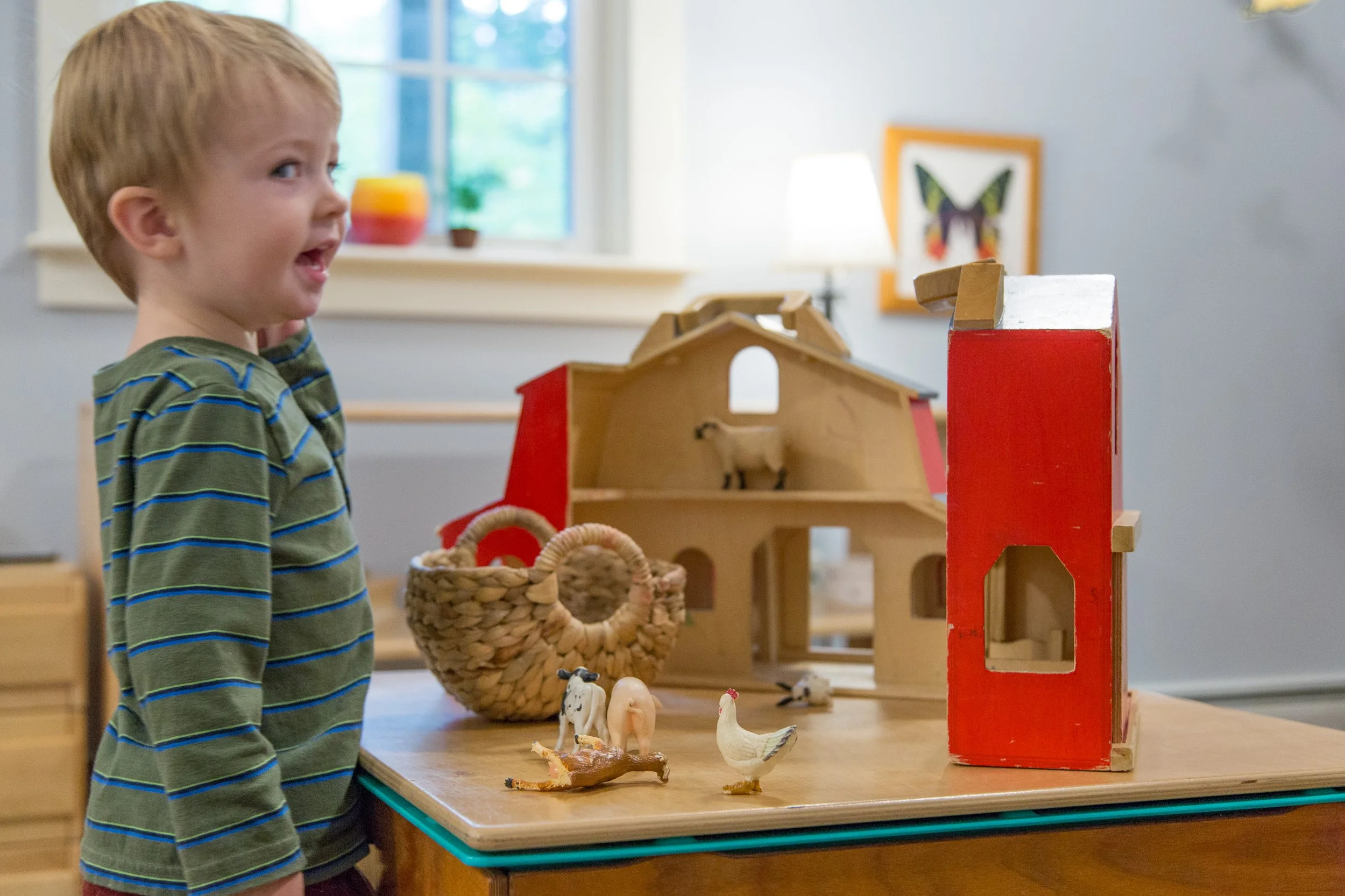 A young boy with blonde hair and a striped green and blue shirt playing with wooden farm toy set, including animals and a barn, on a table in a bright room.