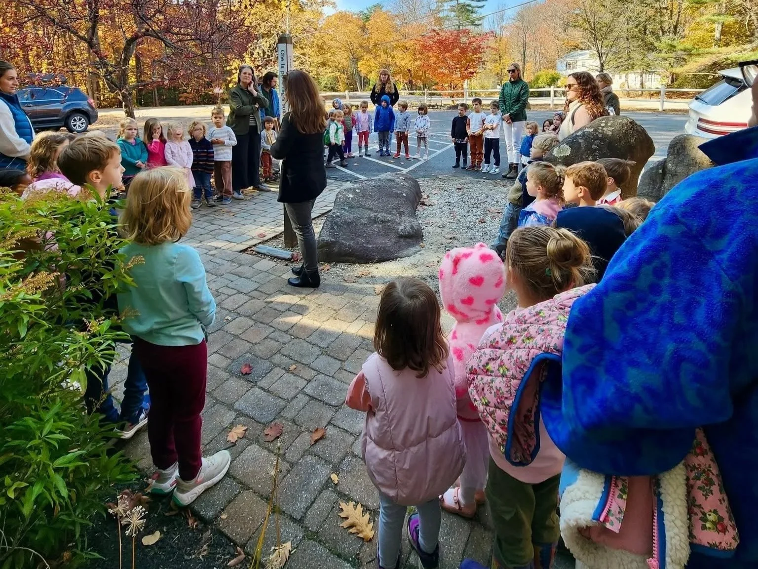 A group of children and adults gathered outdoors in a semicircle around a woman speaking, in a parking lot with autumn trees in the background.