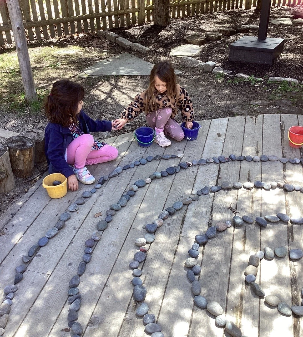 Two young girls playing with rocks on a wooden deck, arranging them in patterns or shapes.