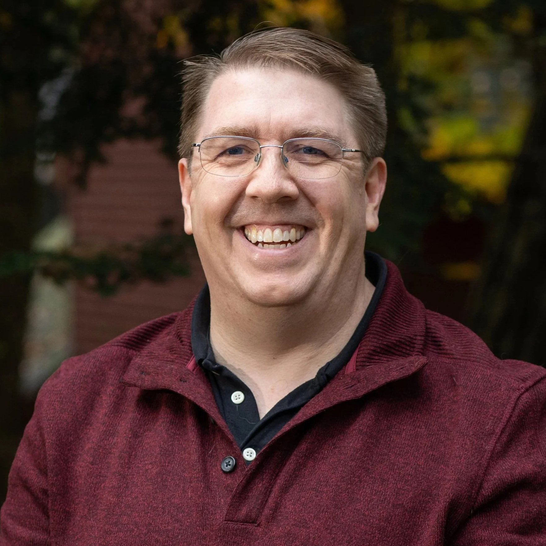 A smiling man wearing glasses and a maroon sweater outdoors with trees and blurred buildings in the background.