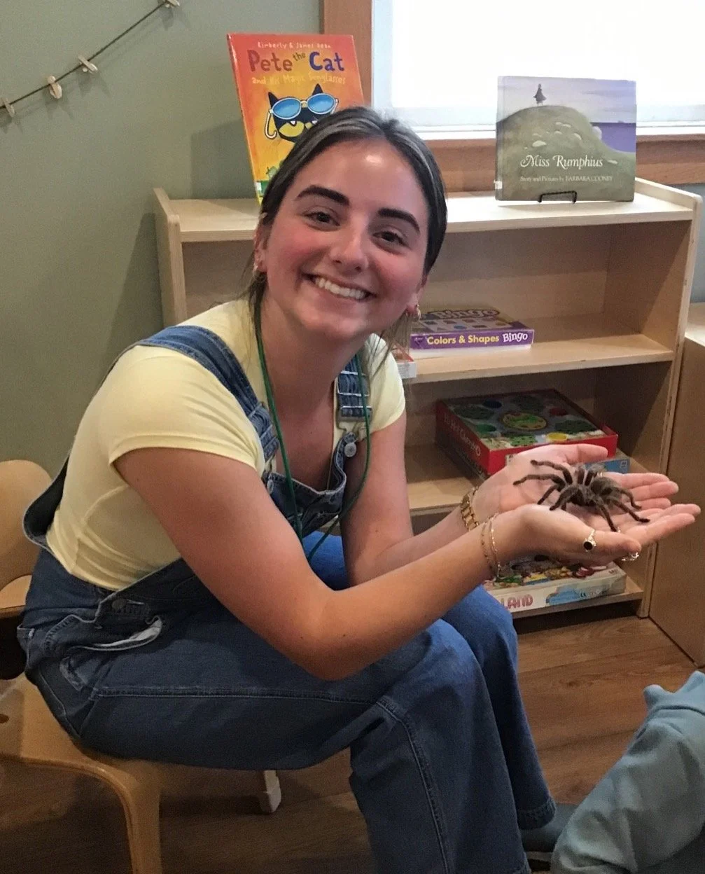 A woman with dark hair, smiling, wearing a yellow shirt and denim overalls, sits on a wooden chair holding a tarantula in her hands in a room with bookshelves and children's books.