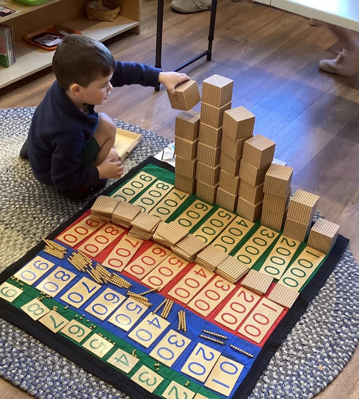 A young boy sitting on a rug playing a Montessori math game with colored number tiles, counters, and a tower of wooden blocks representing numbers.