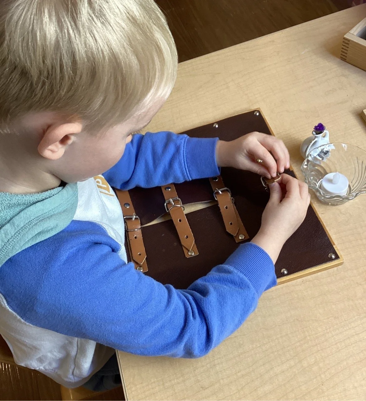 A young boy with blonde hair is sitting at a wooden table, working on a leather craft project. He is using a needle and thread to sew leather straps onto a piece of leather. There is a small decorative item with a purple flower inside a white ceramic pitcher and a glass bowl with a white candle on the table.