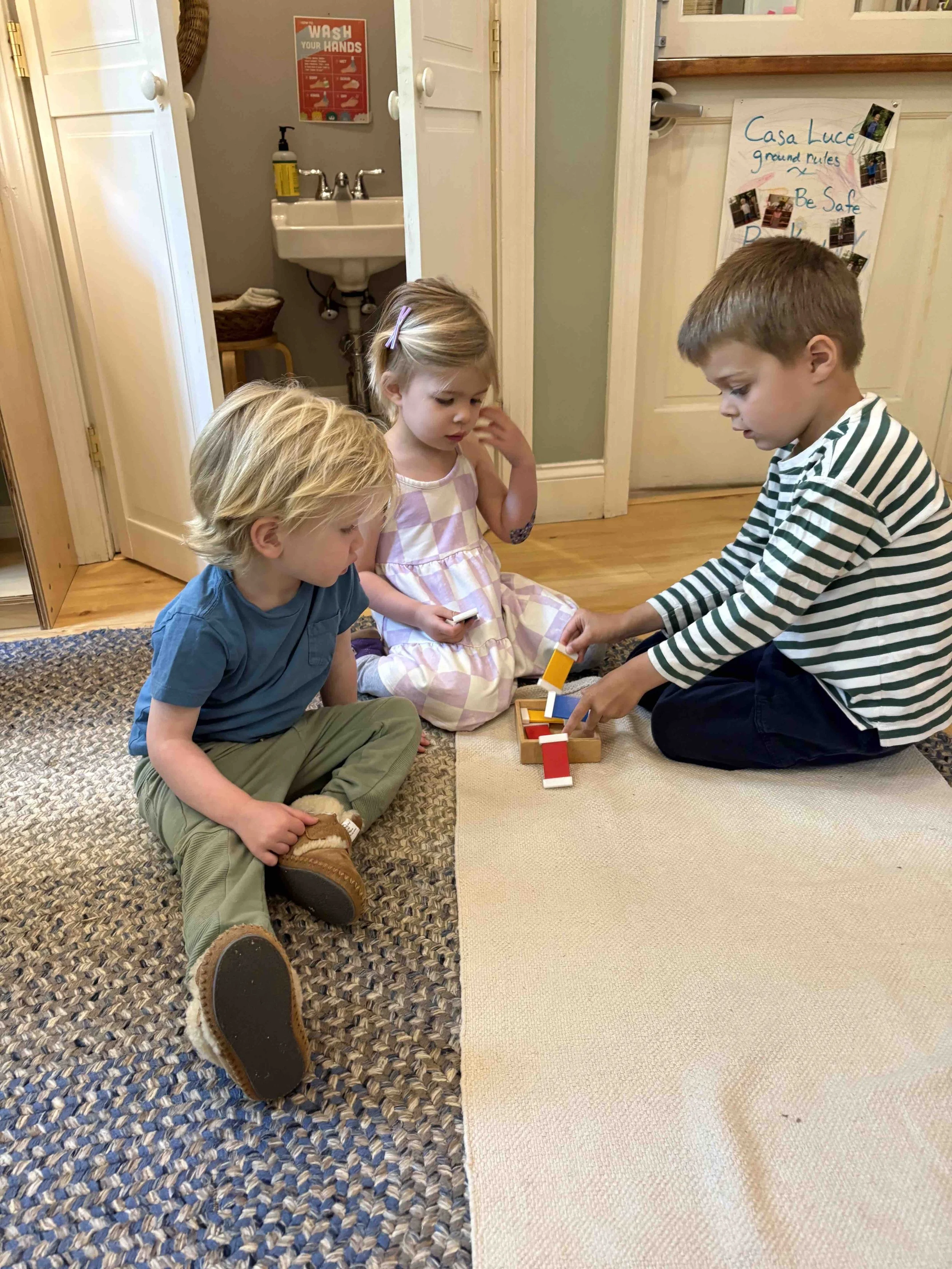 Three children sitting on the floor in a room with beige and carpeted floor, using the Montessori color tablets, with a door and a sink in the background. A sign with handwashing instructions is on the wall.