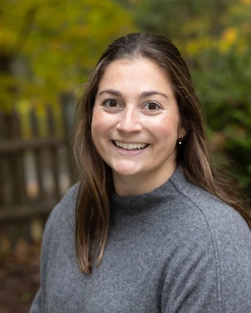 A woman smiling outdoors with a wooden fence and trees with green and yellow leaves in the background.
