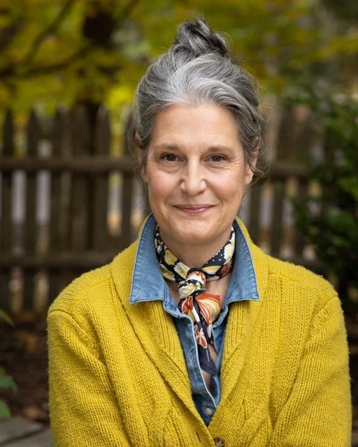 A woman with hair tied up, wearing a yellow cardigan over a denim shirt and a colorful scarf, standing outdoors with a wooden fence and green foliage in the background.