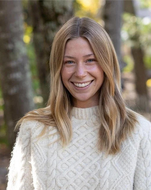 A woman with blonde hair and a big smile, wearing a cream-colored knit sweater, outdoors with trees in the background.
