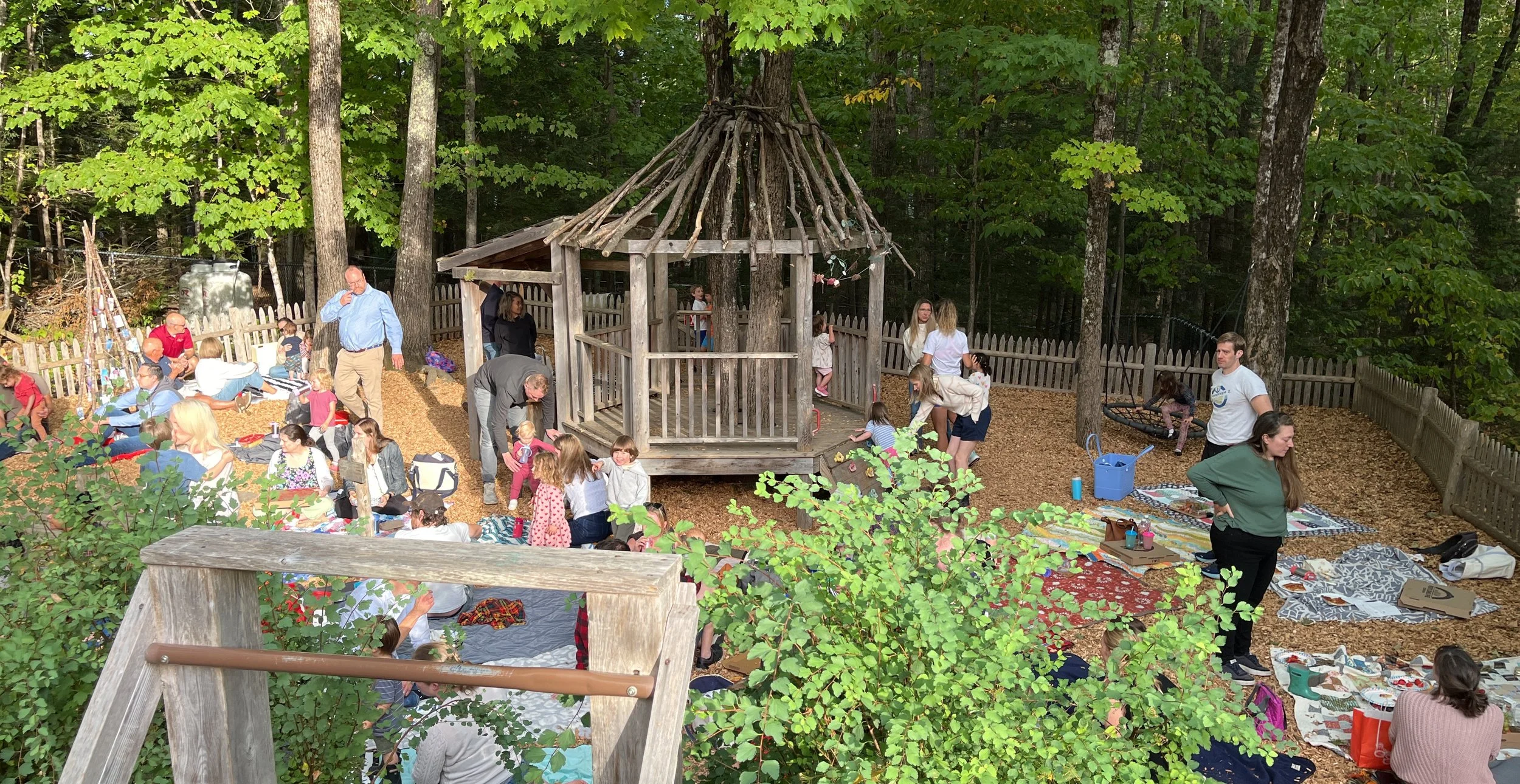 People gather in a wooded outdoor area with a small wooden gazebo, surrounded by trees and fencing, for a social event or gathering.