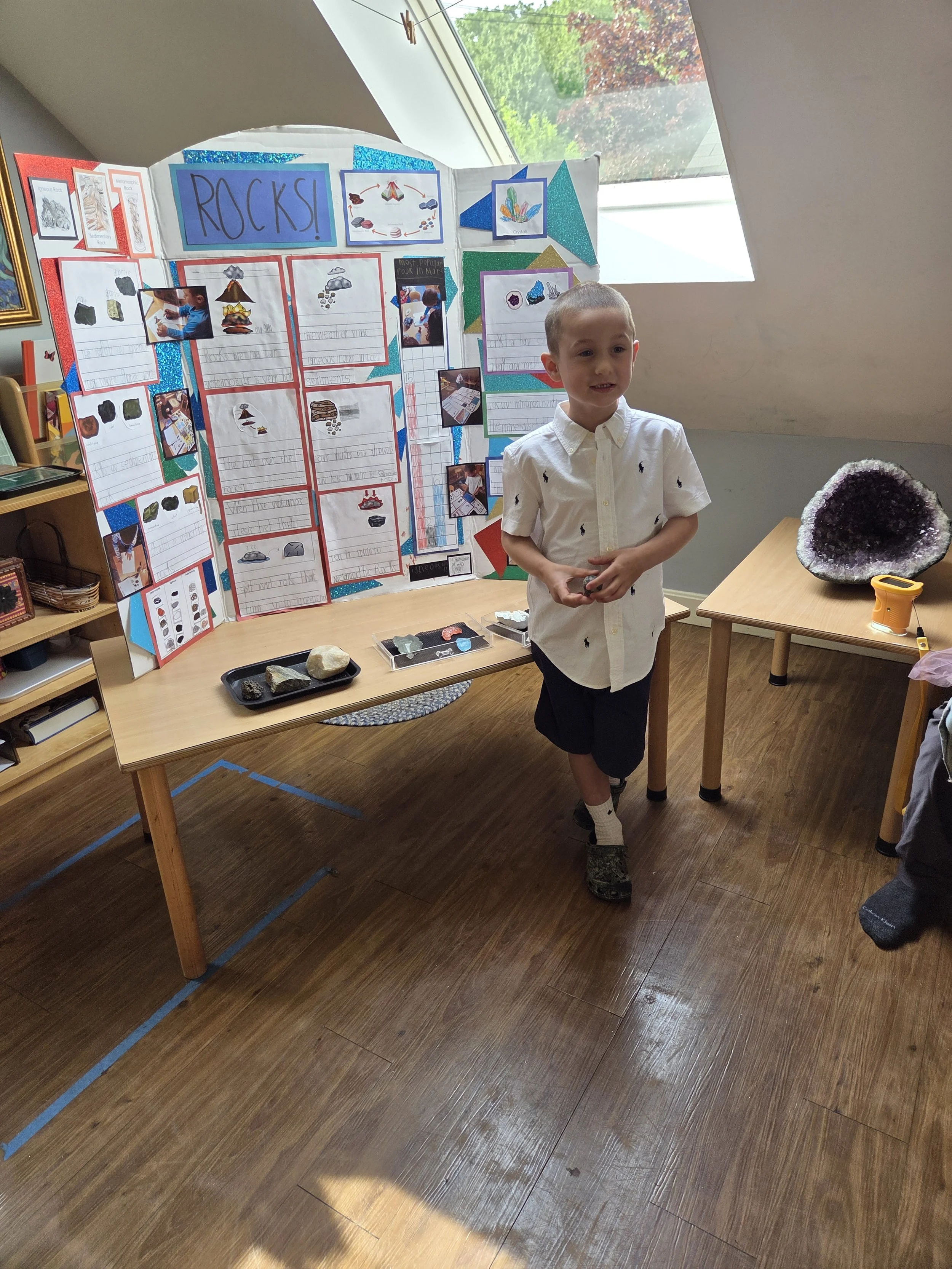 A young boy stands in front of a science project display about rocks, featuring pictures, descriptive text, and various rocks on a table. A large amethyst geode is on a separate table nearby, and sunlight is coming through a window.