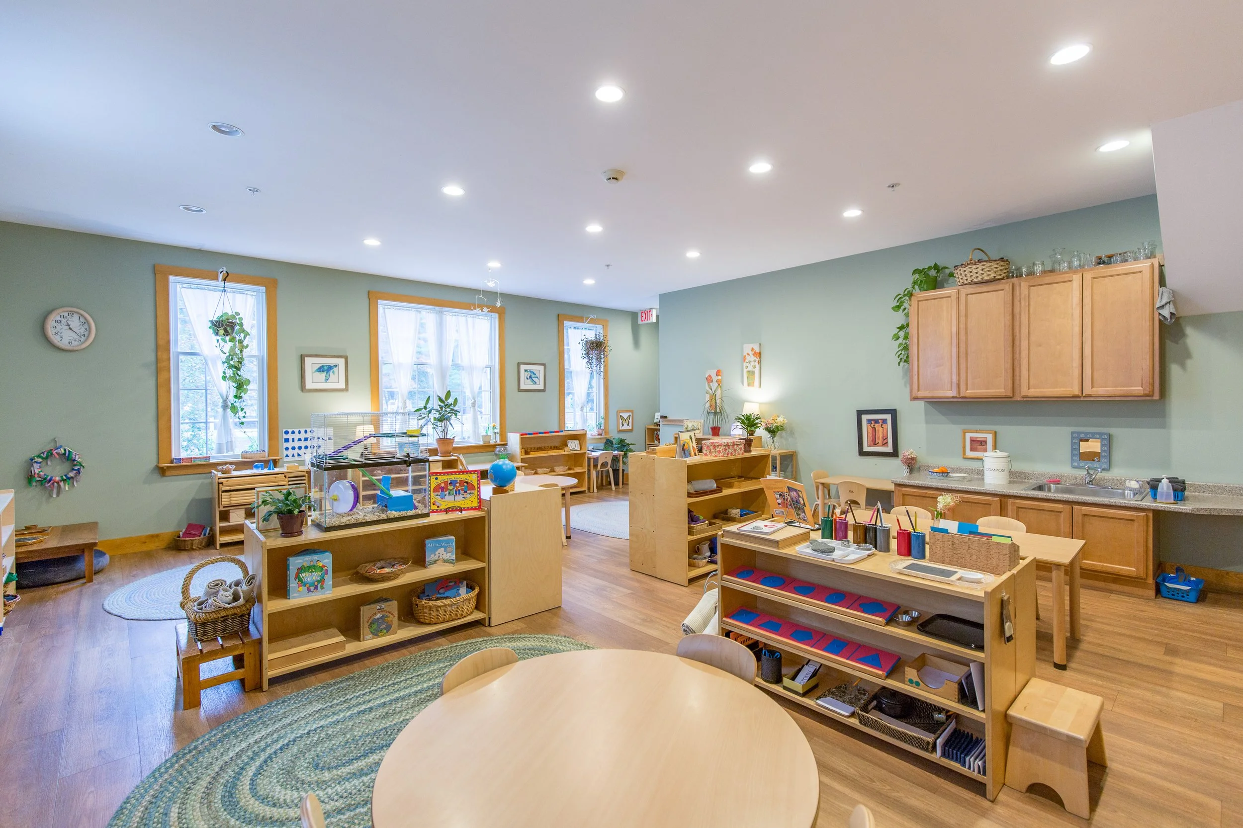 Bright, organized preschool classroom with wooden furniture, educational toys, plants, and colorful decorations.
