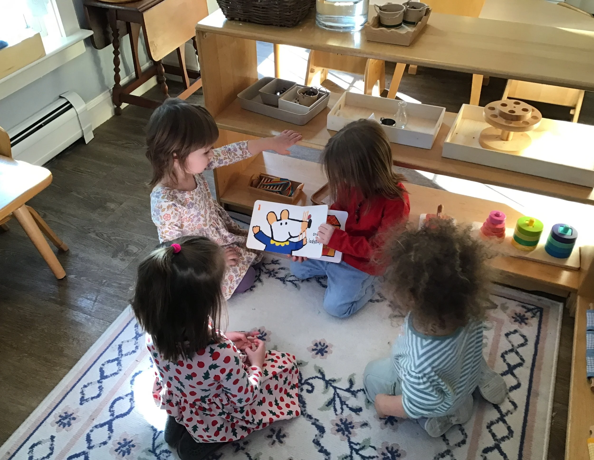 Children sitting on a rug, playing with toys, in a room with wooden furniture and a bench with trays of small objects and craft supplies.