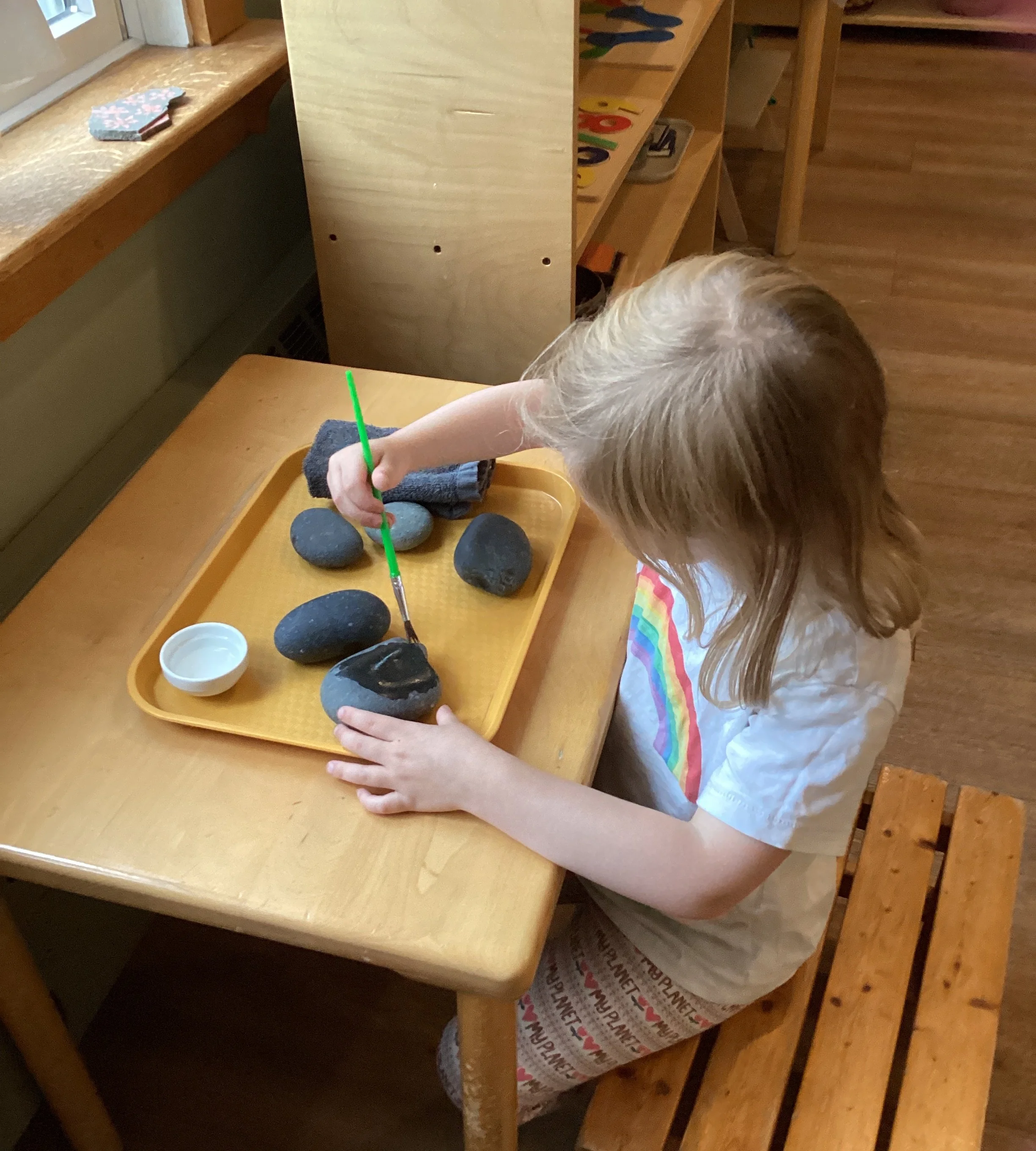 Child with blonde hair completing a craft project at a small wooden table, using a green stick to paint or decorate oval-shaped black stones, with a tray of stones and a small round container nearby, in a room with wooden furniture.