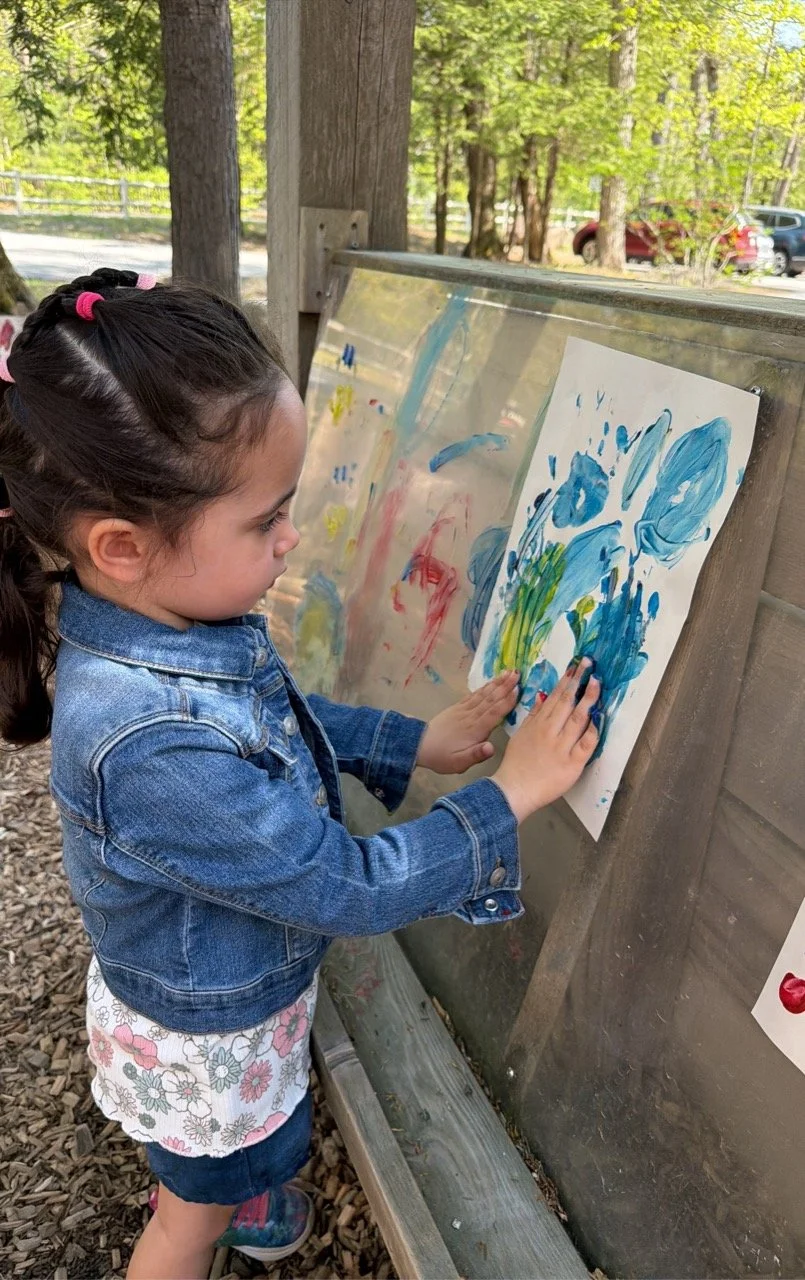 Young girl with a denim jacket and floral skirt painting with blue, red, and yellow colors on a piece of paper attached to an outdoor easel.