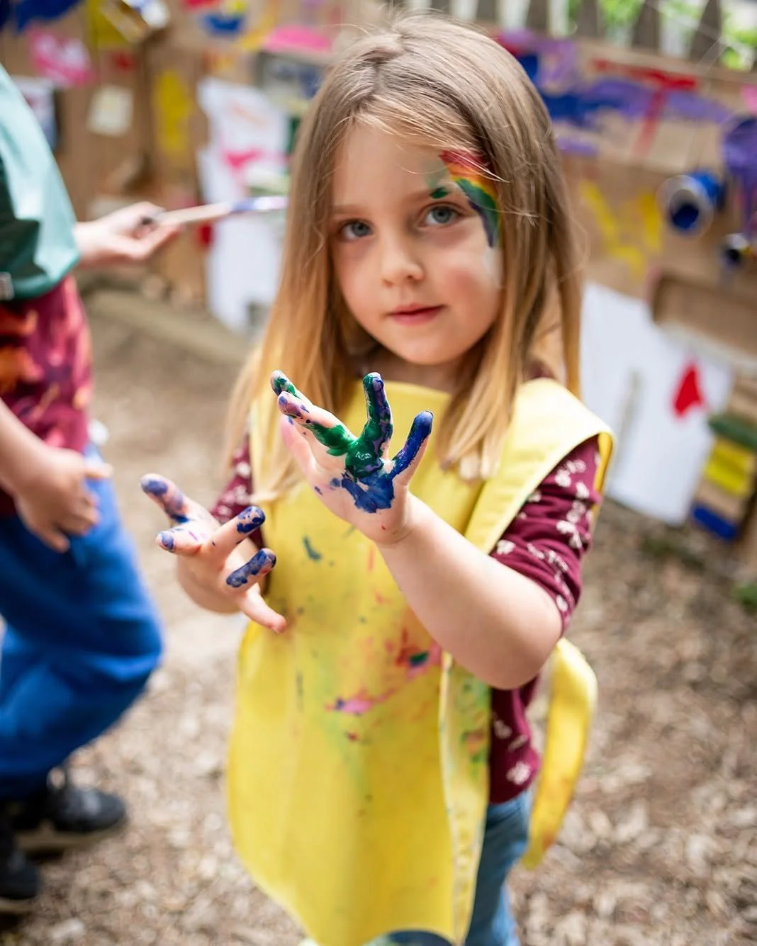 A young girl with paint on her face and hands, showing a 'peace' sign with her fingers, standing outdoors near a painting station.