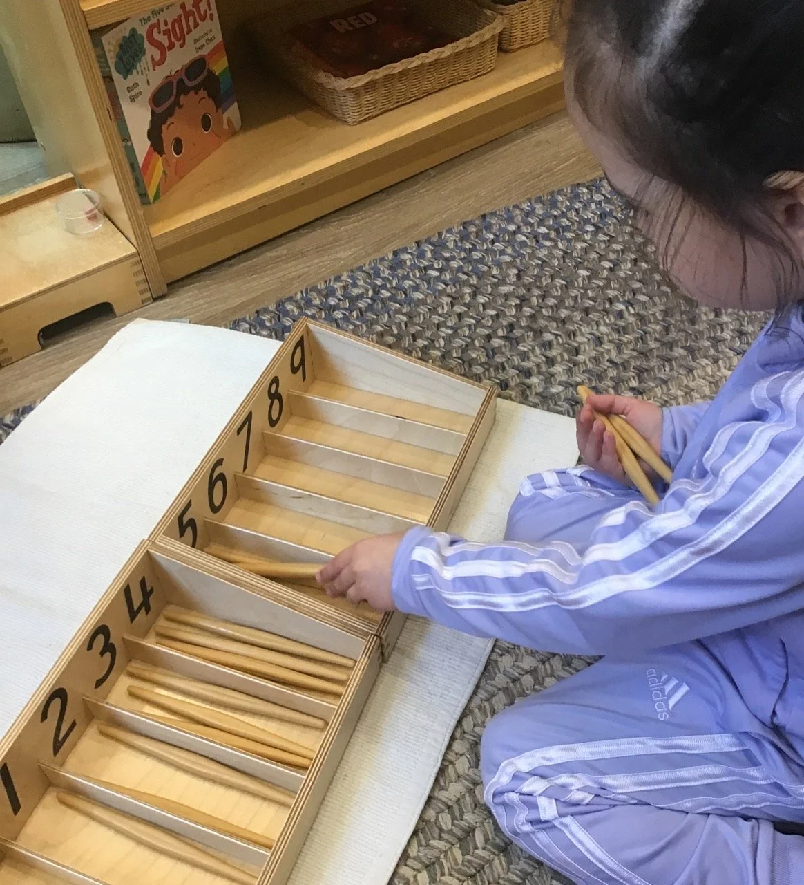 A young girl in a light purple Adidas tracksuit sitting on a beige textured rug, holding chopsticks, and playing with a wooden number tray with numbered compartments from 1 to 9.