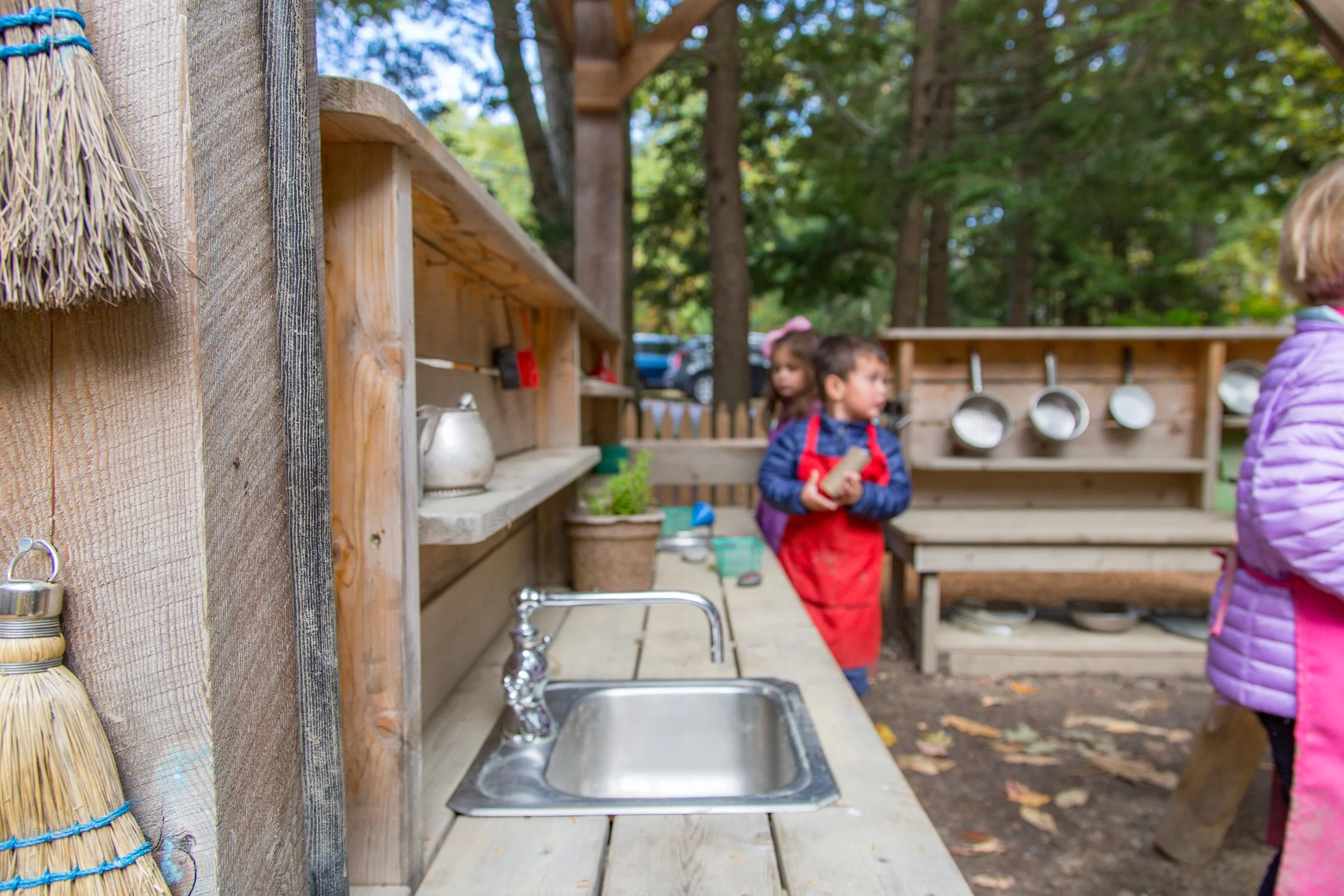 Children playing outdoors in a rustic outdoor kitchen area with wooden shelves and utensils, surrounded by trees.