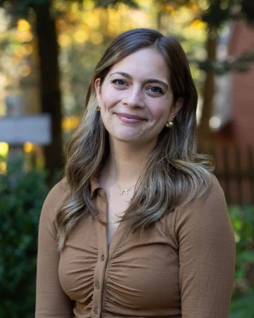A woman with long wavy hair, wearing a brown top, smiling outdoors with trees and a wooden fence in the background.