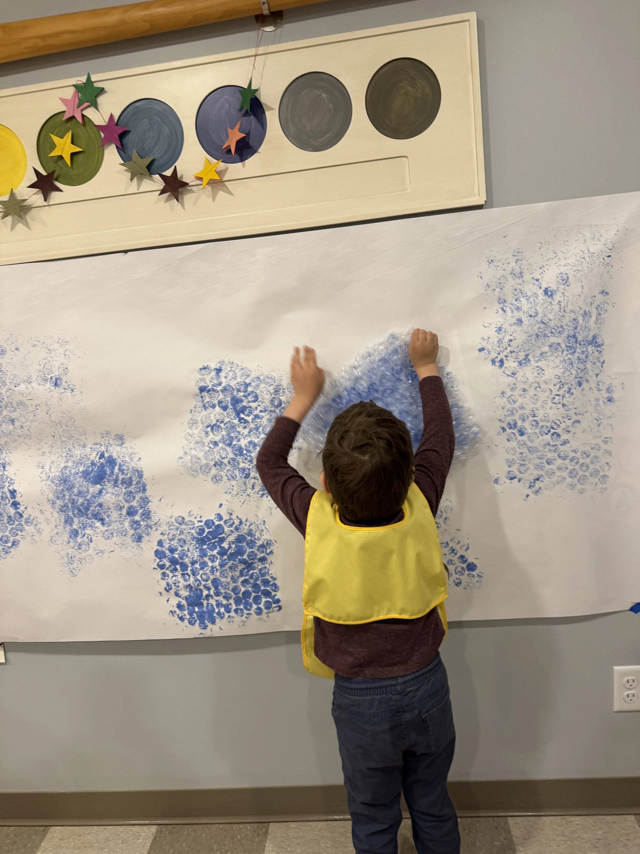 A child wearing a yellow apron is creating art with blue paint on a large white paper wall.