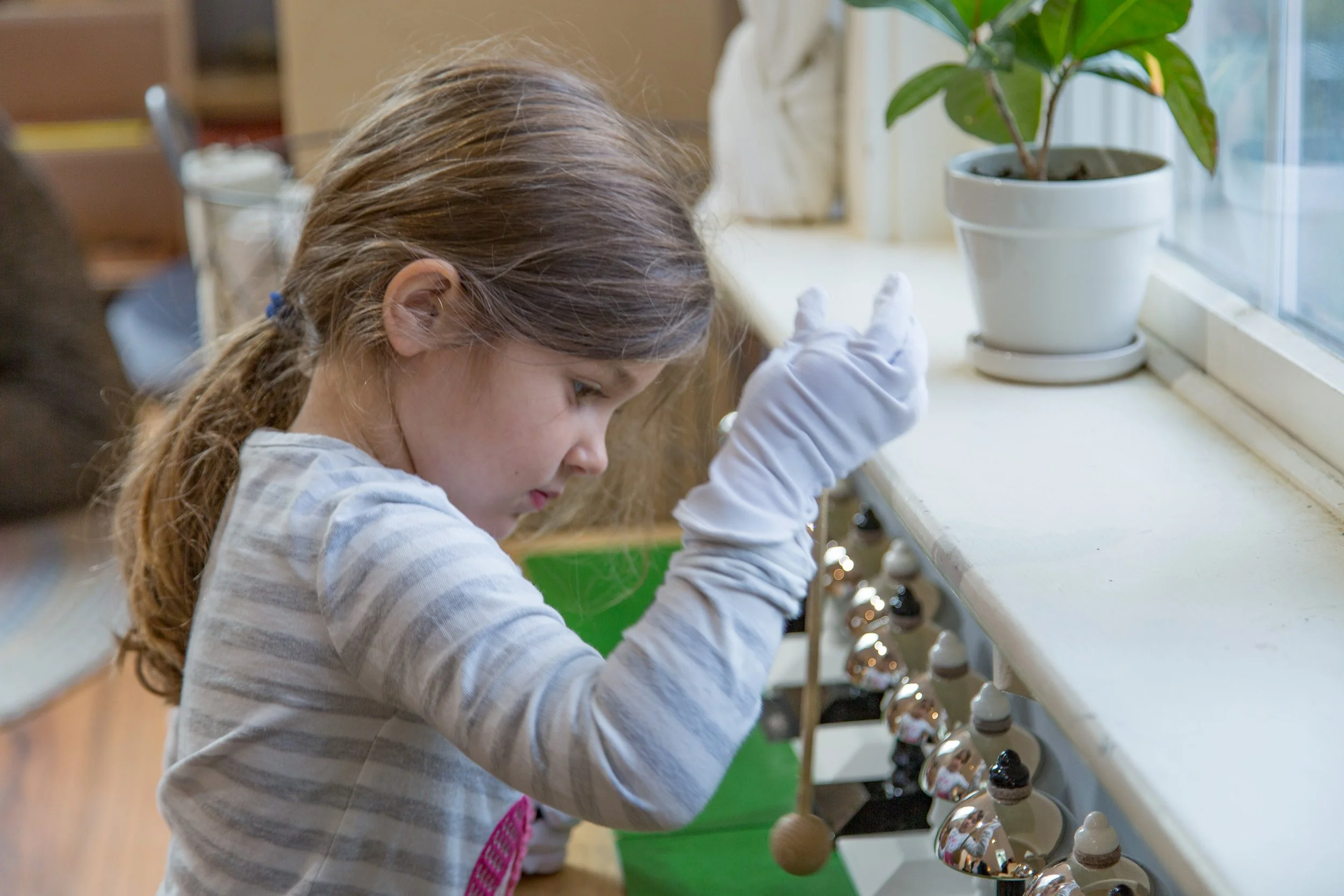 A young girl wearing a gray striped shirt and white gloves is cleaning bell-shaped objects, possibly holiday ornaments, at a white shelf by a window with a potted plant nearby.