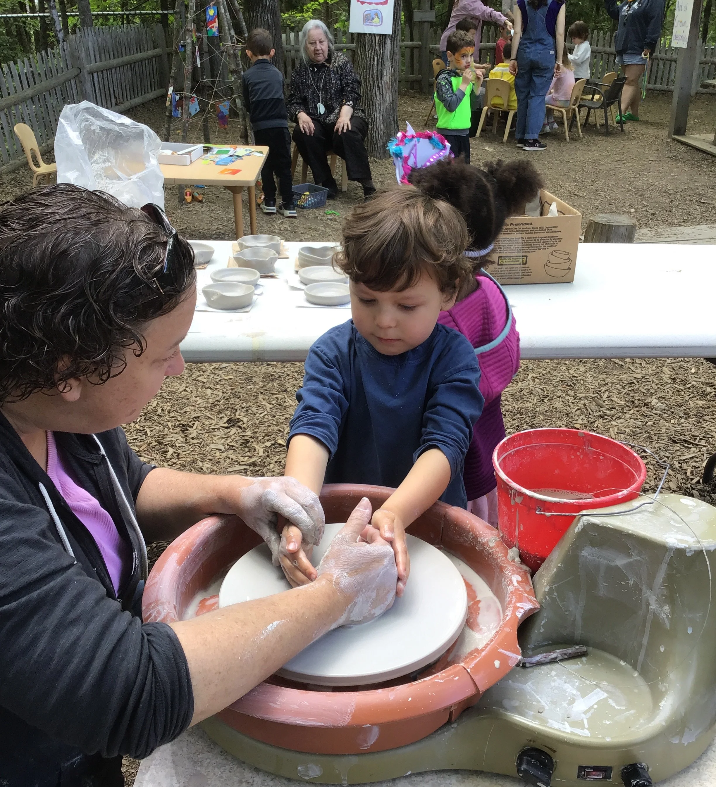A woman helping a young boy learn pottery on a spinning wheel outdoors, with a girl watching. Other children and adults are in the background at a children's event or festival.