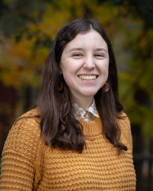 A young woman with long brown hair smiling outdoors, wearing a mustard yellow sweater and a white collared shirt with a floral pattern, with autumn trees in the background.