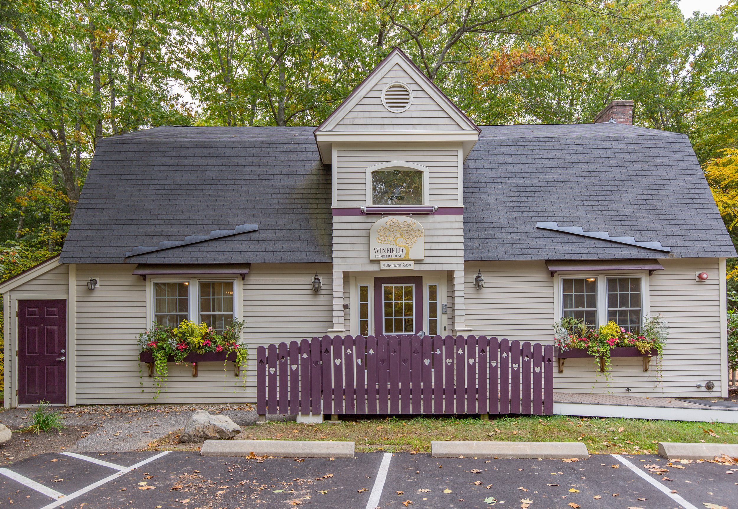 A small, two-story building with beige siding and a dark gray shingled roof. The building has two windows with flower boxes, purple trim and purple fencing in front, and a sign reading "Winfield Toddler House" above the purple door. Trees with green leaves are in the background.