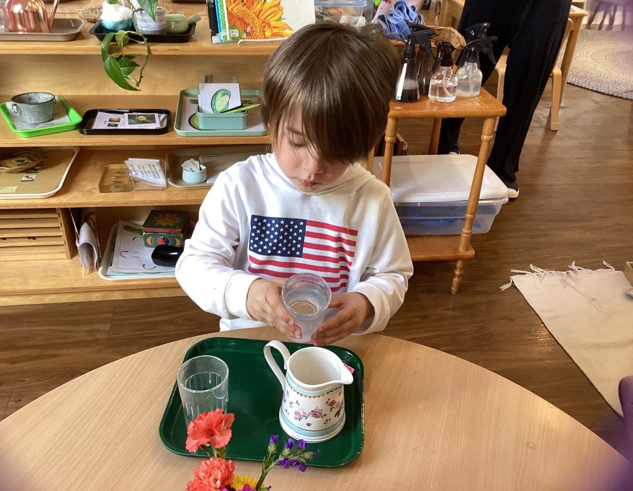 A young boy with brown hair, wearing a white hoodie with an American flag on it, is pouring water from a glass into a pitcher on a green tray. The tray also has an empty glass and a floral-patterned creamer. There are colorful flowers in a vase in th