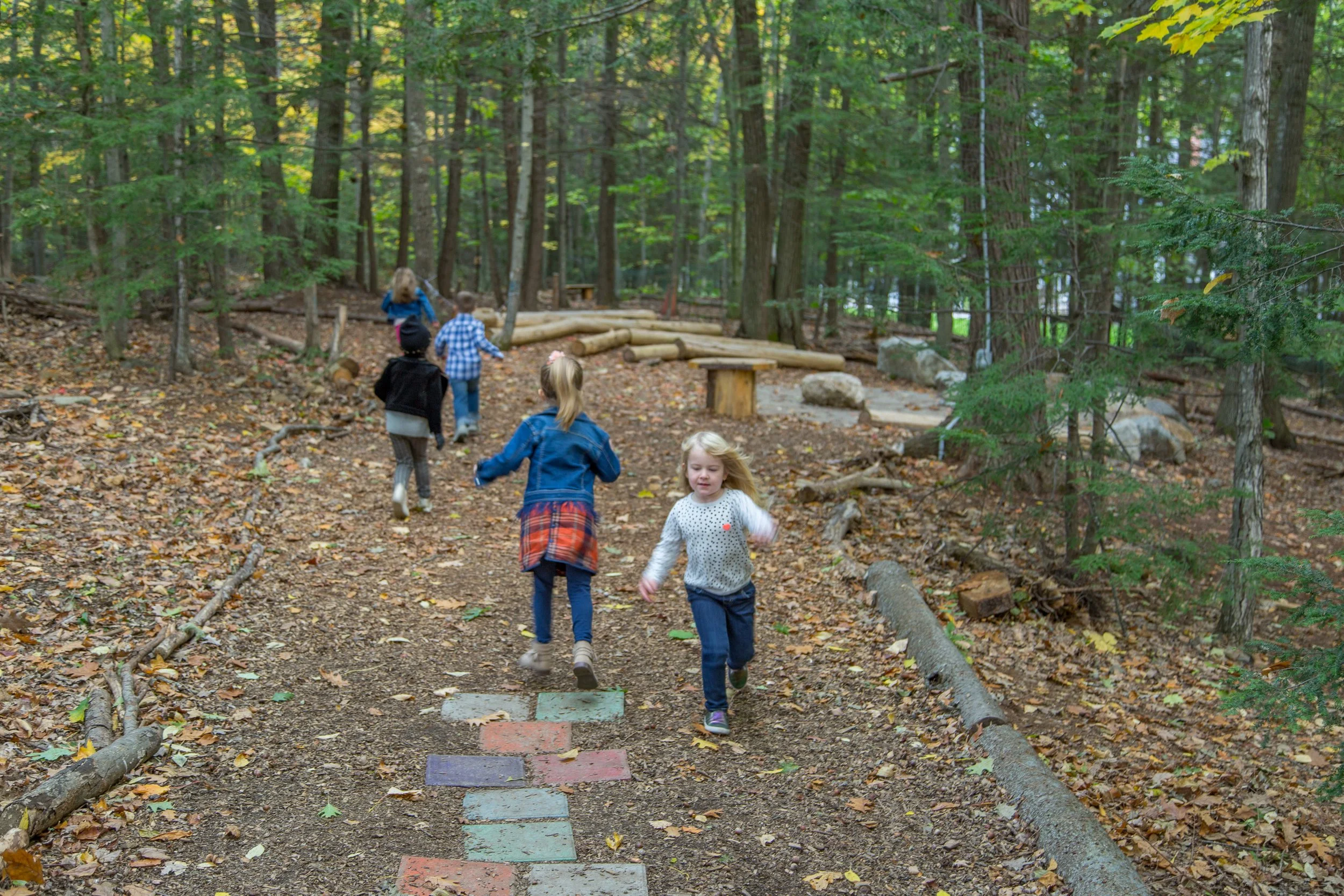 Children playing and running in an outdoor classroom with trees, leaves, and a dirt path.
