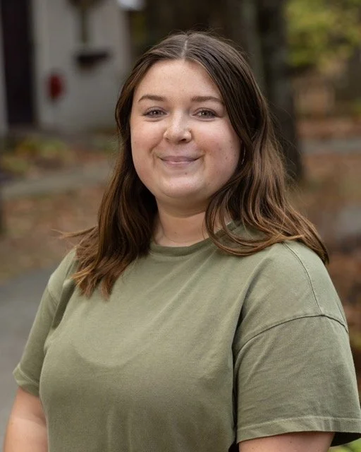 A young woman with brown hair and a nose piercing smiling outdoors, wearing a green t-shirt.