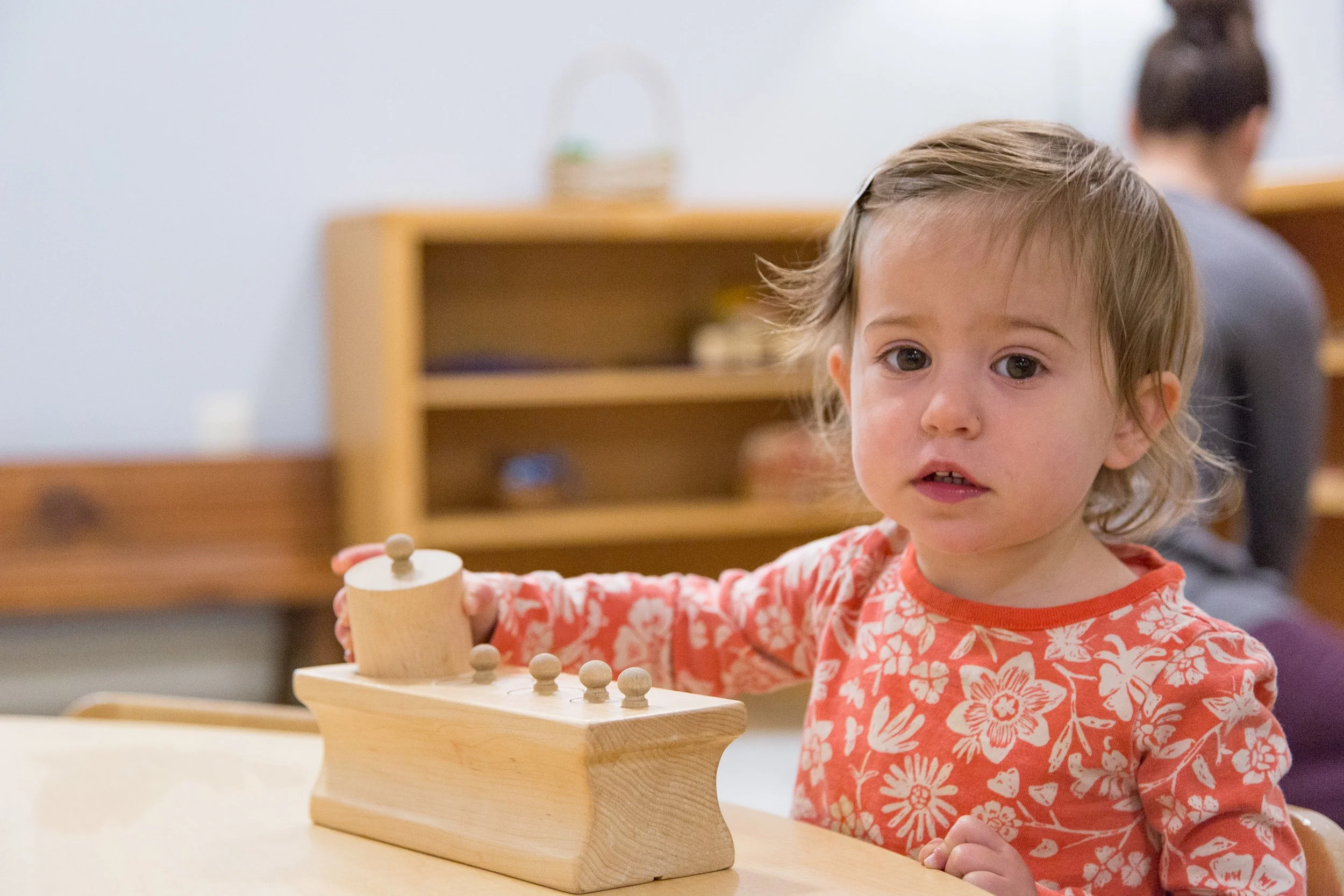 Young girl with light brown hair and wearing a orange and white floral shirt playing with a wooden toy on a table, with a woman in a gray shirt blurred in the background.